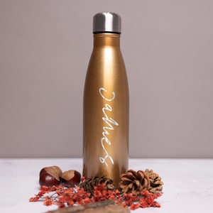 Könnte beinhalten: Eine goldene Wasserflasche aus Metall mit einer weißen Aufschrift "Christmas". Die Flasche steht auf einer weißen Oberfläche mit Tannenzapfen und roten Beeren.