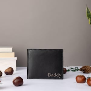 May include: A black bi-fold wallet featuring the word Daddy embossed in the bottom right corner. The wallet has a smooth, matte finish and a rectangular shape, standing upright on a flat surface with visible stitching along the edges.
