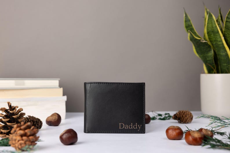 May include: A black bi-fold wallet featuring the word Daddy embossed in the bottom right corner. The wallet has a smooth, matte finish and a rectangular shape, standing upright on a flat surface with visible stitching along the edges.