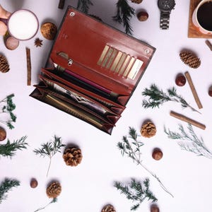 May include: A brown leather wallet with multiple card slots and a zippered compartment is open and lying on a white surface. The wallet is surrounded by small pine cones, twigs, and other natural elements.
