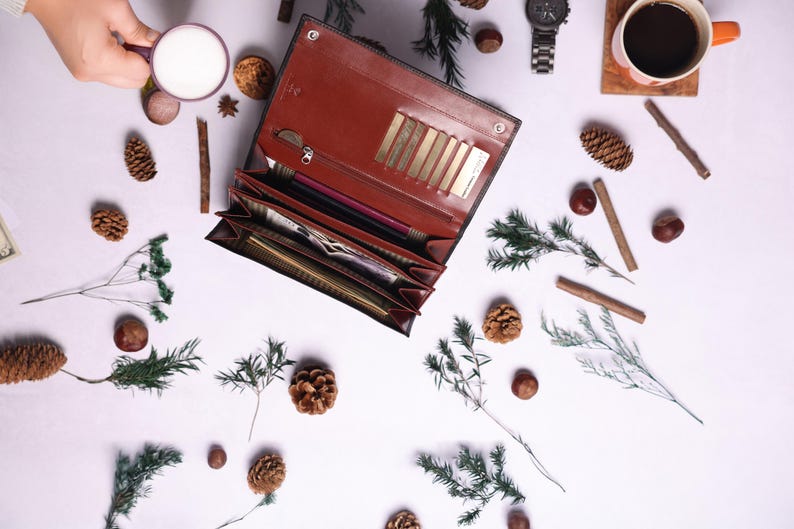May include: A brown leather wallet with multiple card slots and a zippered compartment is open and lying on a white surface. The wallet is surrounded by small pine cones, twigs, and other natural elements.