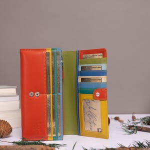 May include: A multi-colored leather wallet featuring a vibrant orange exterior, a lime green interior, and various card slots in shades of yellow, teal, and blue. The wallet includes a clear ID window, a snap button closure, and visible stitching.