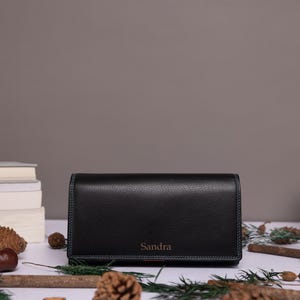 May include: A black rectangular wallet with a pebbled texture and visible stitching along the edges. The name Sandra is embossed in a small, centered font on the front flap of the wallet. The wallet is positioned horizontally on a flat surface.