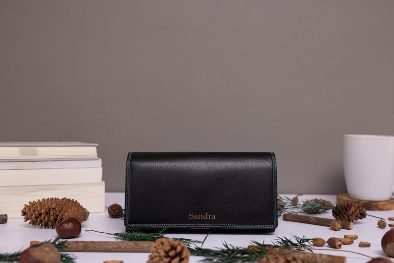May include: A black rectangular wallet with a pebbled texture and visible stitching along the edges. The name Sandra is embossed in a small, centered font on the front flap of the wallet. The wallet is positioned horizontally on a flat surface.