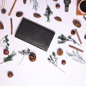 May include: A flat lay image of a black leather wallet, a cup of coffee, a watch, and various natural elements, including pine cones, cinnamon sticks, and sprigs of greenery.