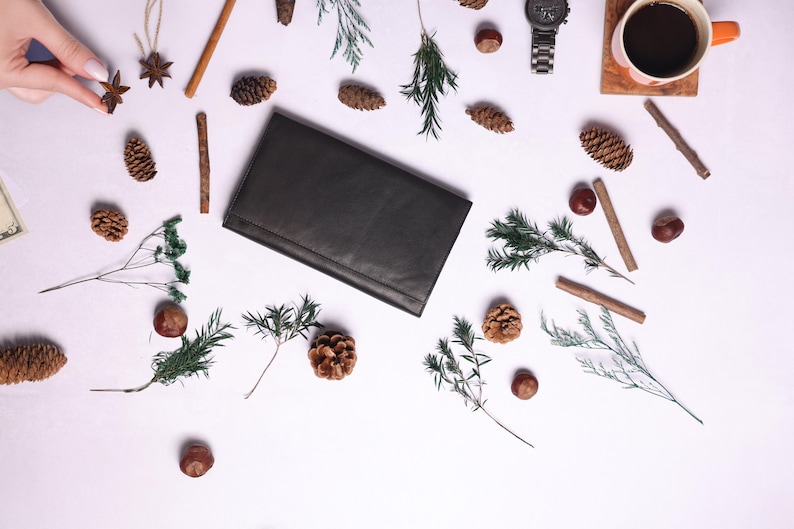 May include: A flat lay image of a black leather wallet, a cup of coffee, a watch, and various natural elements, including pine cones, cinnamon sticks, and sprigs of greenery.
