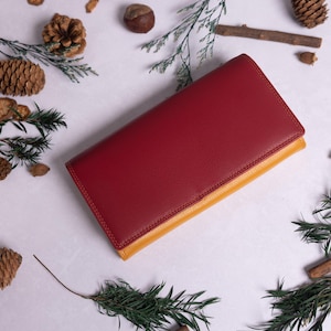 May include: A red leather wallet with a yellow leather trim. The wallet is lying on a white surface with pine cones and branches scattered around it.