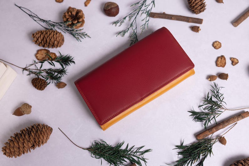 May include: A red leather wallet with a yellow leather trim. The wallet is lying on a white surface with pine cones and branches scattered around it.