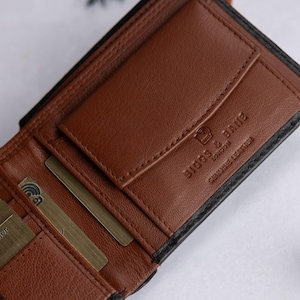 May include: A brown leather bifold wallet with a black exterior. The interior features multiple card slots, a coin pocket with a flap, and a plaid fabric lining. The wallet is embossed with the text BIGGS & BANE LONDON and GENUINE LEATHER.