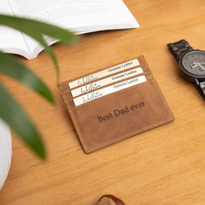 May include: A brown leather cardholder with the text "Best Dad ever" is displayed on a wooden surface. Three cards are visible in the cardholder. A black watch with a dark face and a wooden band is next to the cardholder.