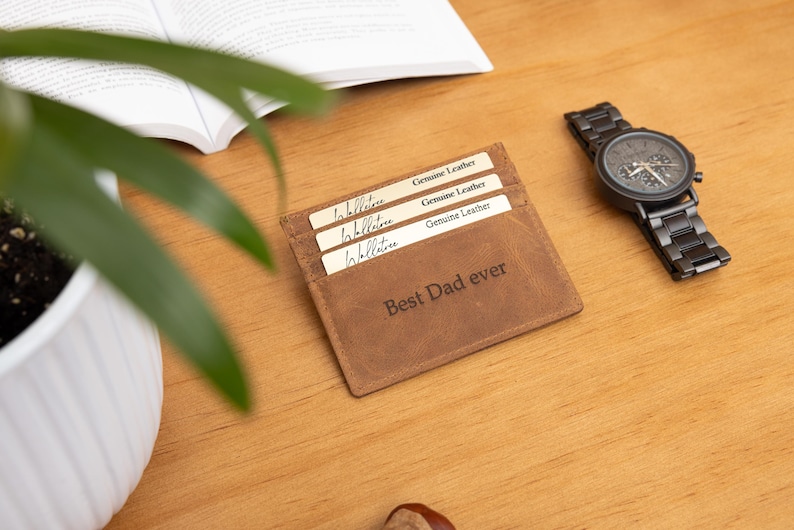 May include: A brown leather cardholder with the text "Best Dad ever" is displayed on a wooden surface. Three cards are visible in the cardholder. A black watch with a dark face and a wooden band is next to the cardholder.