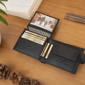 May include: Black leather wallet with multiple card slots and a photo insert. The wallet is open, revealing a picture and several gold-colored cards. A black watch with a dark face is next to the wallet. The wallet is on a wooden surface.
