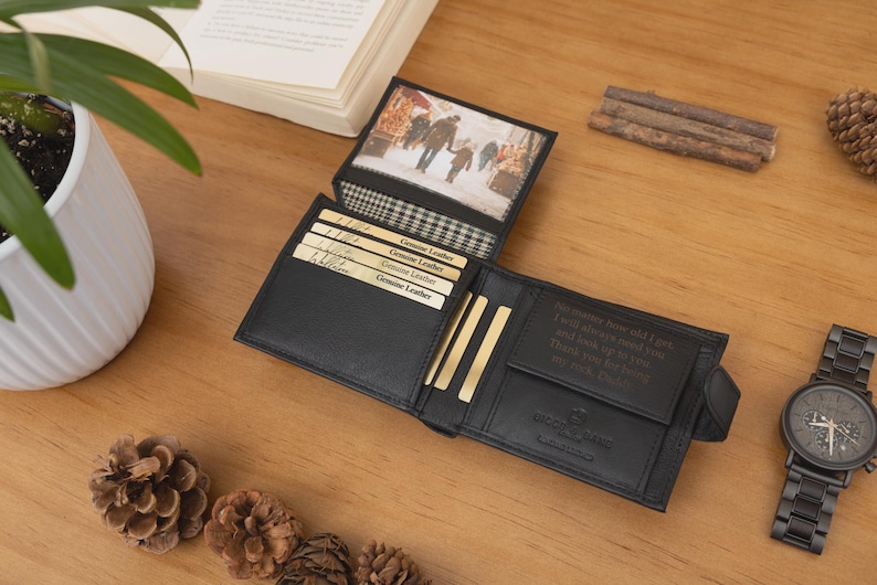 May include: Black leather wallet with multiple card slots and a photo insert. The wallet is open, revealing a picture and several gold-colored cards. A black watch with a dark face is next to the wallet. The wallet is on a wooden surface.