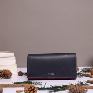 May include: A rectangular navy blue wallet with a pebbled texture and the name Millie embossed in gold lettering on the front flap. The wallet features a contrasting dark red interior lining visible along the bottom edge of the closed accessory.