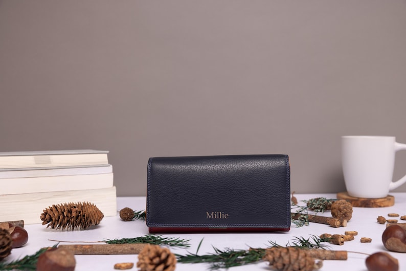 May include: A rectangular navy blue wallet with a pebbled texture and the name Millie embossed in gold lettering on the front flap. The wallet features a contrasting dark red interior lining visible along the bottom edge of the closed accessory.