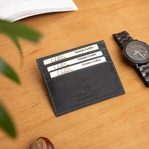 May include: A black leather cardholder with the word "Dad" embossed on the front, next to a black wristwatch with a metal band. The cardholder contains three white cards that read "Genuine Leather". The items are on a wooden surface.