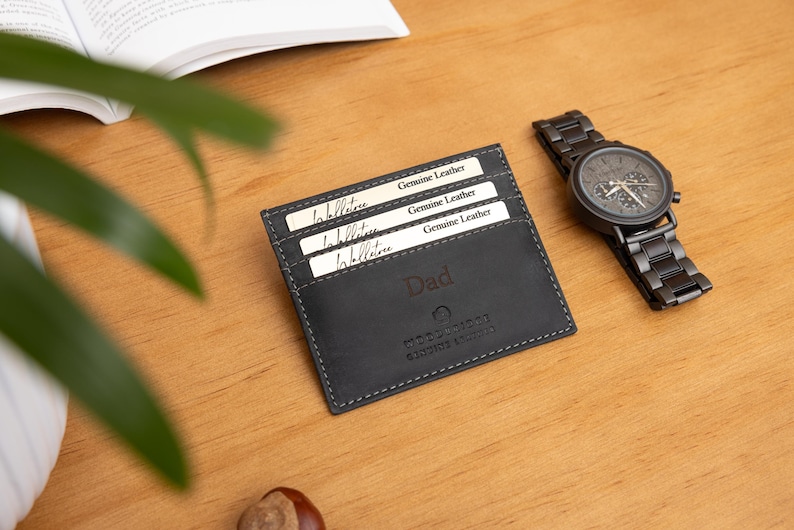 May include: A black leather cardholder with the word "Dad" embossed on the front, next to a black wristwatch with a metal band. The cardholder contains three white cards that read "Genuine Leather". The items are on a wooden surface.