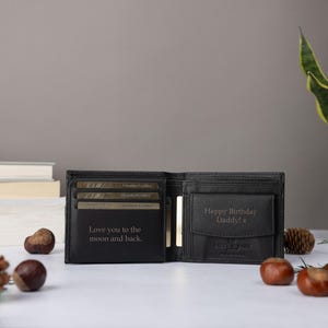 May include: A black bi-fold wallet featuring interior engravings. The left side displays the text "Love you to the moon and back." The right side features a coin pocket with the engraved text "Happy Birthday Daddy! x" and a small logo.