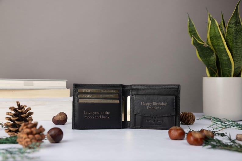 May include: A black bi-fold wallet featuring interior engravings. The left side displays the text "Love you to the moon and back." The right side features a coin pocket with the engraved text "Happy Birthday Daddy! x" and a small logo.
