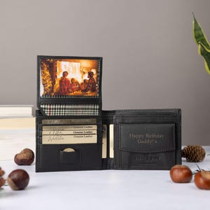 May include: A black leather bi-fold wallet featuring a vertical flip-up photo insert displaying a family scene, a plaid fabric lining, multiple card slots, and a coin pocket embossed with the text Happy Birthday Daddy! x in a dark font.