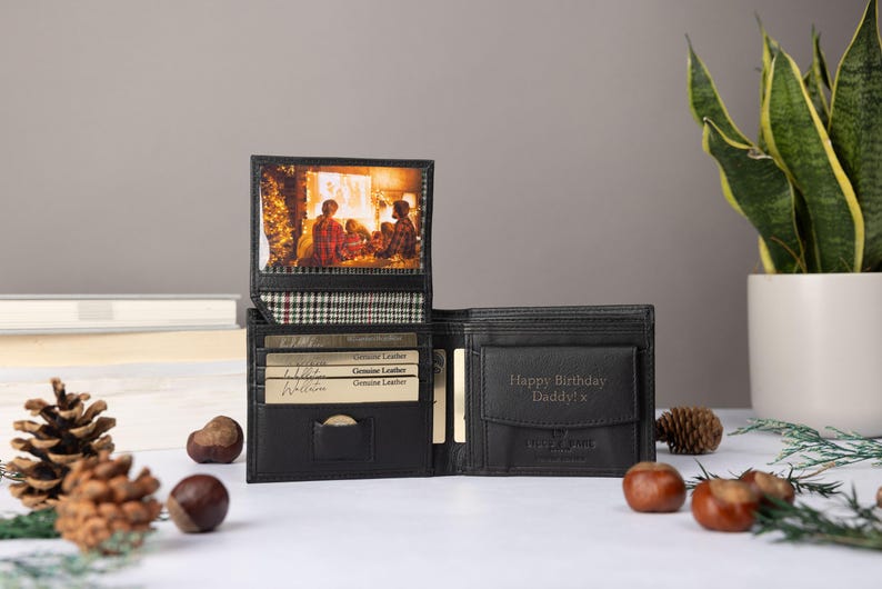 May include: A black leather bi-fold wallet featuring a vertical flip-up photo insert displaying a family scene, a plaid fabric lining, multiple card slots, and a coin pocket embossed with the text Happy Birthday Daddy! x in a dark font.