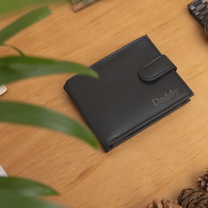 May include: Black leather wallet with the word "Daddy" engraved on the front. A black wristwatch with a matching band is next to the wallet. The items are on a wooden surface, with greenery and pine cones in the background.