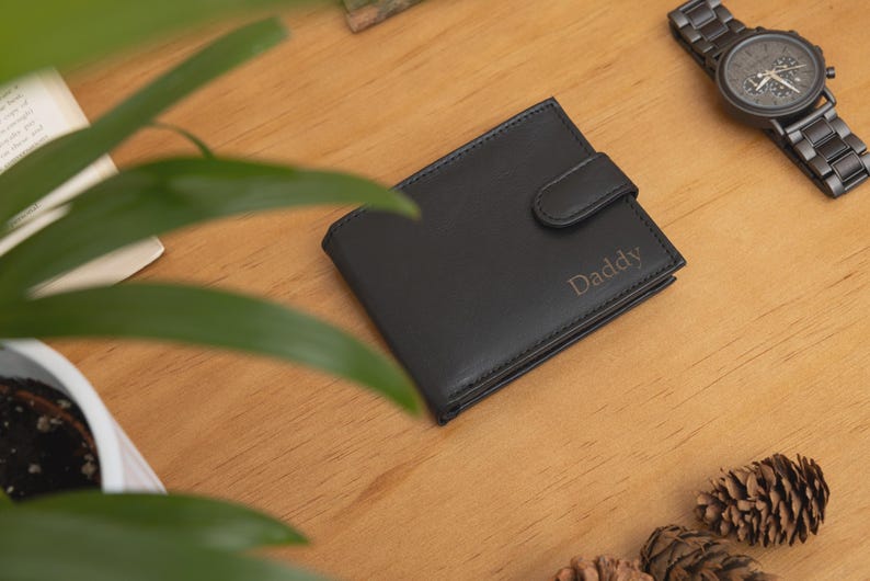 May include: Black leather wallet with the word "Daddy" engraved on the front. A black wristwatch with a matching band is next to the wallet. The items are on a wooden surface, with greenery and pine cones in the background.