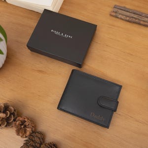 May include: Black leather wallet with the word "Daddy" engraved on the front, next to a black watch and a black box. The box has the words "Biggs & Bane" printed on it. The items are on a wooden surface.