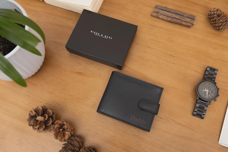May include: Black leather wallet with the word "Daddy" engraved on the front, next to a black watch and a black box. The box has the words "Biggs & Bane" printed on it. The items are on a wooden surface.