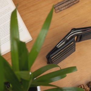 May include: An open black leather wallet with visible cards and cash sits on a wooden surface. A partially visible open book, a wristwatch, and a small plant are also on the table. Pine cones and small wooden sticks are scattered around.