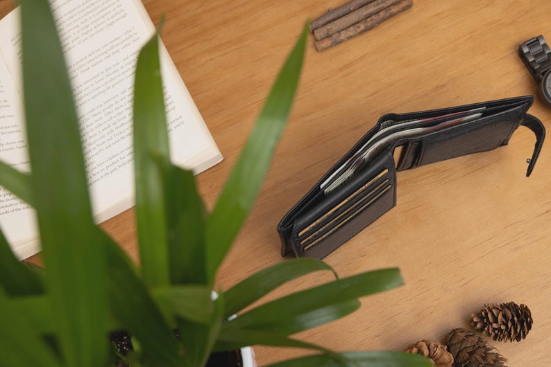 May include: An open black leather wallet with visible cards and cash sits on a wooden surface. A partially visible open book, a wristwatch, and a small plant are also on the table. Pine cones and small wooden sticks are scattered around.