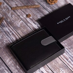 Może przedstawiać: Czarny sk&oacute;rzany portfel z szarym klapką i srebrnym tłoczonym logo z napisem "BIGGS & DANE DESIGN LONDON" w czarnym pudełku.