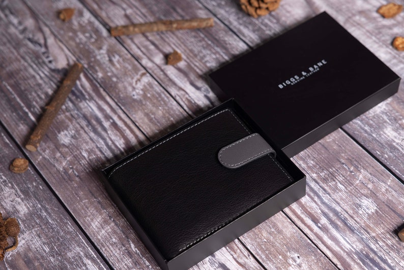 Może przedstawiać: Czarny sk&oacute;rzany portfel z szarym klapką i srebrnym tłoczonym logo z napisem "BIGGS & DANE DESIGN LONDON" w czarnym pudełku.