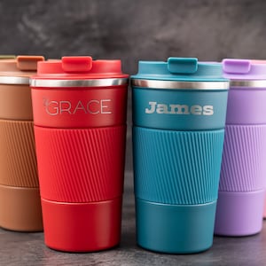 May include: Seven personalized travel mugs in various colors, including red, green, blue, purple, and white. The mugs have names engraved on them, including "Grace" and "James".