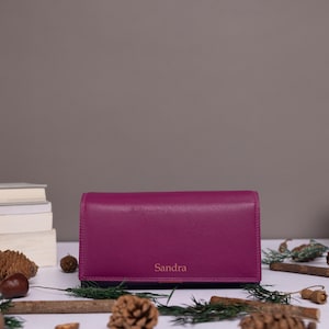 May include: A rectangular magenta leather wallet featuring the name Sandra embossed in a simple font on the front flap. The wallet is positioned horizontally with a smooth, matte finish and clean, straight edges, showcasing a solid color design.