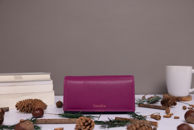 May include: A rectangular magenta leather wallet featuring the name Sandra embossed in a simple font on the front flap. The wallet is positioned horizontally with a smooth, matte finish and clean, straight edges, showcasing a solid color design.