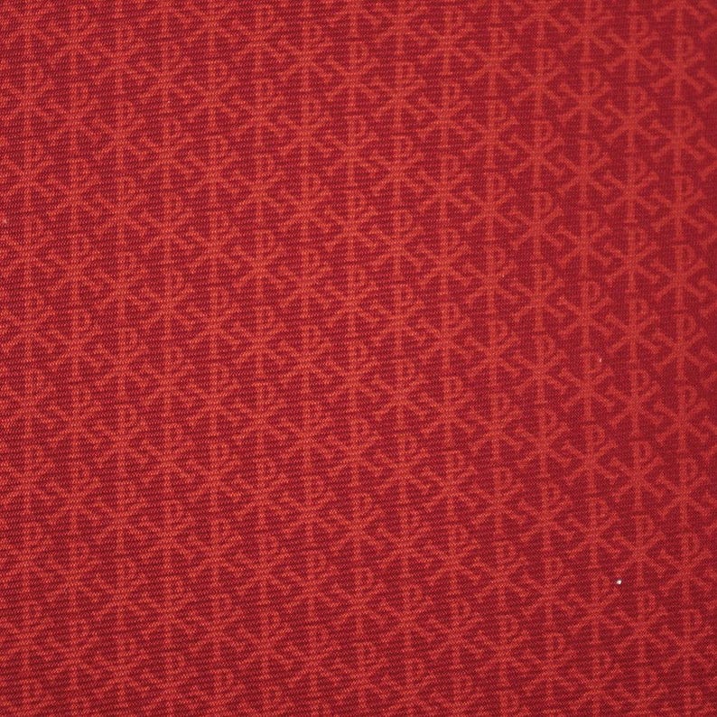 May include: Close-up of a red fabric with a repeating pattern of stylized symbols. The symbols are a lighter shade of red, creating a subtle contrast. The fabric has a textured appearance.