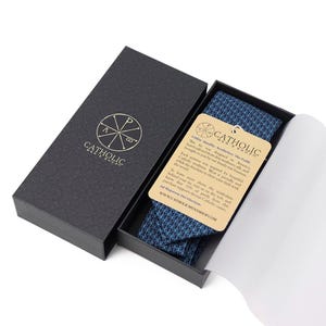 May include: A navy blue patterned tie presented in a black box. The box is open, revealing the tie and a tag with text. The tie has a geometric design. The box has the words "Catholic Mens Shop" and a logo.