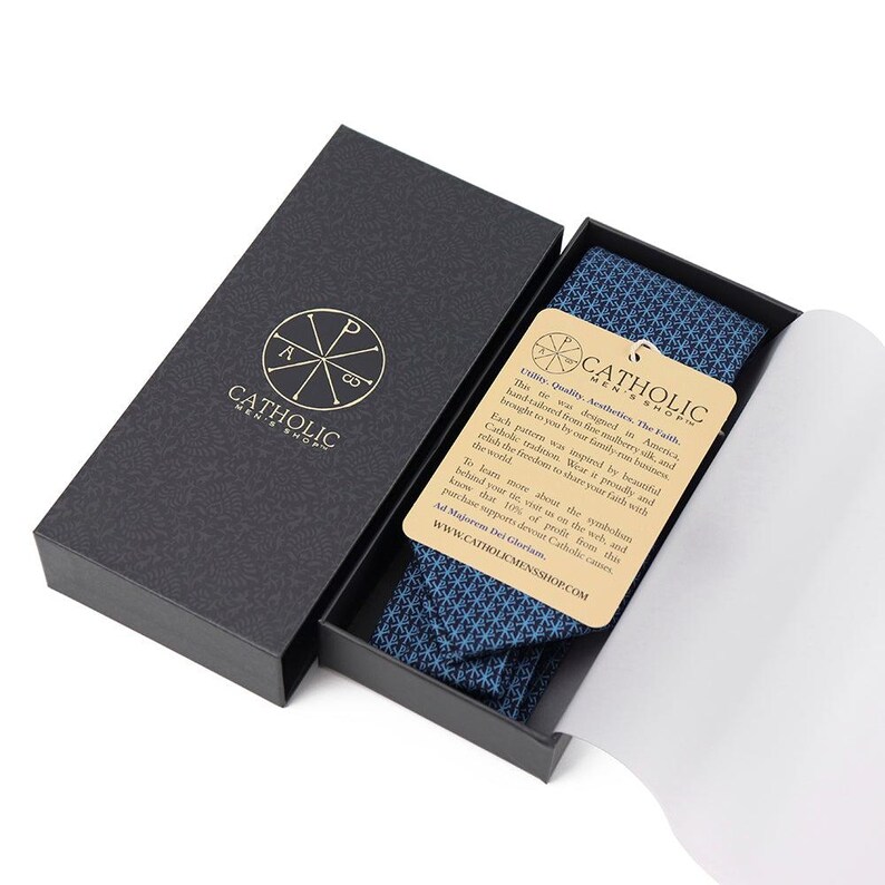 May include: A navy blue patterned tie presented in a black box. The box is open, revealing the tie and a tag with text. The tie has a geometric design. The box has the words "Catholic Mens Shop" and a logo.