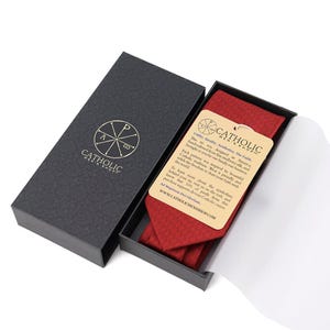 May include: A red tie with a textured pattern is displayed in an open black box. The box has a gold logo and the words "Catholic Men's Shop" printed on it. A tag with text is attached to the tie.