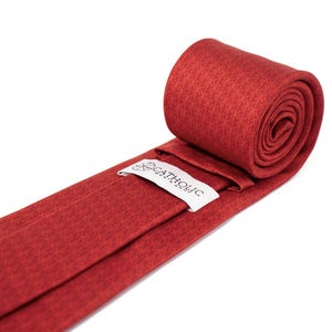 May include: Rolled red tie with a subtle geometric pattern. The tie features a white label with the text "CATHOLIC MENS SHOP". The tie is made of a smooth fabric and is partially rolled up.