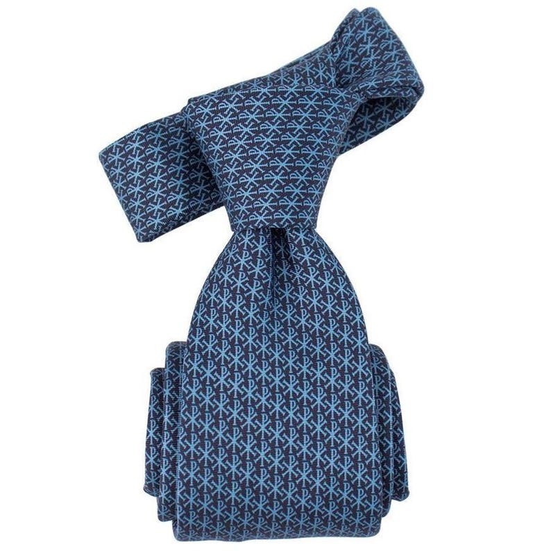 May include: A navy blue tie with a repeating light blue pattern. The pattern consists of a stylized cross and the Greek letters Chi and Rho. The tie is knotted and folded, showcasing the intricate design.