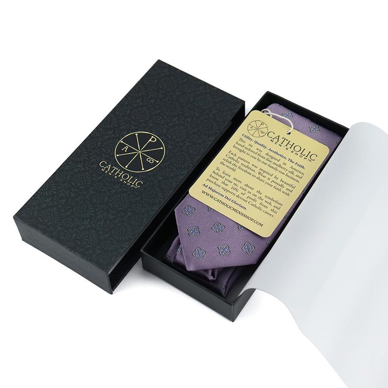May include: A purple silk tie with a white and blue pattern of four-leaf clovers. The tie comes in a black box with a gold logo that says "Catholic Mens Shop". The box also includes a card with the text "Uniting Quality, Authenticity, The Faith. This tie was designed in America, hand-tailored from five-ply mulberry silk, and brought to you by our family business. Each piece was crafted with pride and symbolizes the freedom to share your faith with the world. To learn more about the symbolism behind your tie, visit us on the web and know that 10% of profits from this purchase support Catholic causes. Ad Majorem Dei Gloriam. www.CATHOLICMENSSHOP.COM"