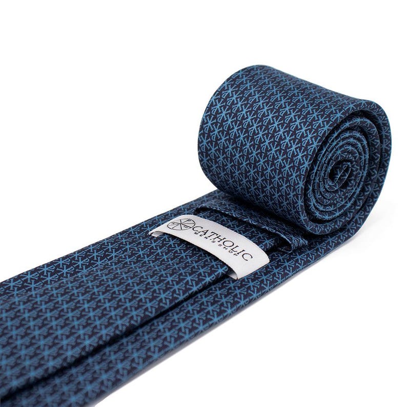 May include: Rolled navy blue tie with a repeating light blue pattern. The tie is partially unrolled, revealing a white tag with the text "CATHOLIC". The tie is made of a smooth fabric.