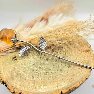 May include: A decorative rose made of amber-colored resin with a silver-colored stem and leaves. The rose is displayed on a wooden slice with dried grasses in the background. The rose is a unique home decor item.