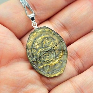 K&ouml;nnte beinhalten: Ein ovaler Anh&auml;nger mit einer goldfarbenen Metalloberfl&auml;che, einem erhabenen Rosenbl&uuml;tenmotiv und sichtbaren horizontalen Rissen, der an einer silberfarbenen Metallkette h&auml;ngt.