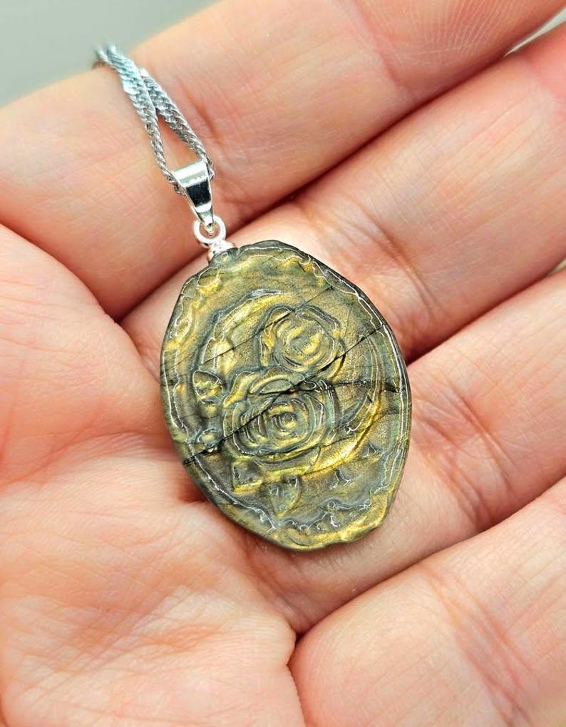 K&ouml;nnte beinhalten: Ein ovaler Anh&auml;nger mit einer goldfarbenen Metalloberfl&auml;che, einem erhabenen Rosenbl&uuml;tenmotiv und sichtbaren horizontalen Rissen, der an einer silberfarbenen Metallkette h&auml;ngt.