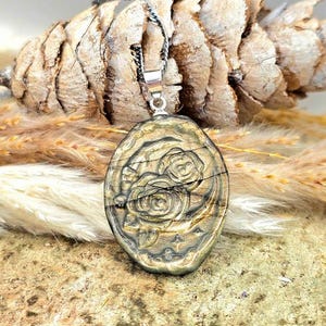 K&ouml;nnte beinhalten: Ein ovaler Anh&auml;nger mit einem geschnitzten Rosenmotiv auf einer dunklen, metallisch wirkenden Steinoberfl&auml;che. Der Anh&auml;nger h&auml;ngt an einer d&uuml;nnen, gedrehten silberfarbenen Kette und zeigt ein detailliertes Blumenrelief mit goldenen Akzenten.