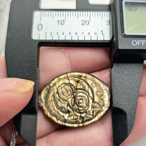 K&ouml;nnte beinhalten: Ein ovaler, goldbrauner Anh&auml;nger mit einem gepr&auml;gten Blumenmuster aus zwei Rosen. Der Anh&auml;nger befindet sich in einer schwarzen digitalen Messschieber-Vorrichtung, die eine Breite von etwa 25 Millimetern an der breitesten Stelle anzeigt.