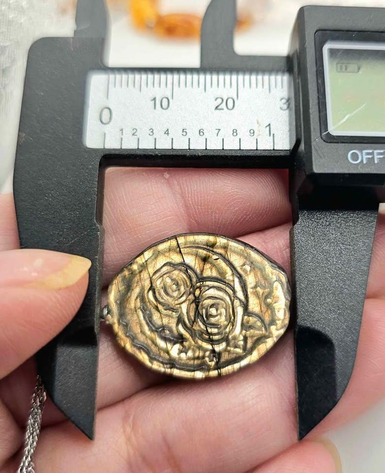 K&ouml;nnte beinhalten: Ein ovaler, goldbrauner Anh&auml;nger mit einem gepr&auml;gten Blumenmuster aus zwei Rosen. Der Anh&auml;nger befindet sich in einer schwarzen digitalen Messschieber-Vorrichtung, die eine Breite von etwa 25 Millimetern an der breitesten Stelle anzeigt.